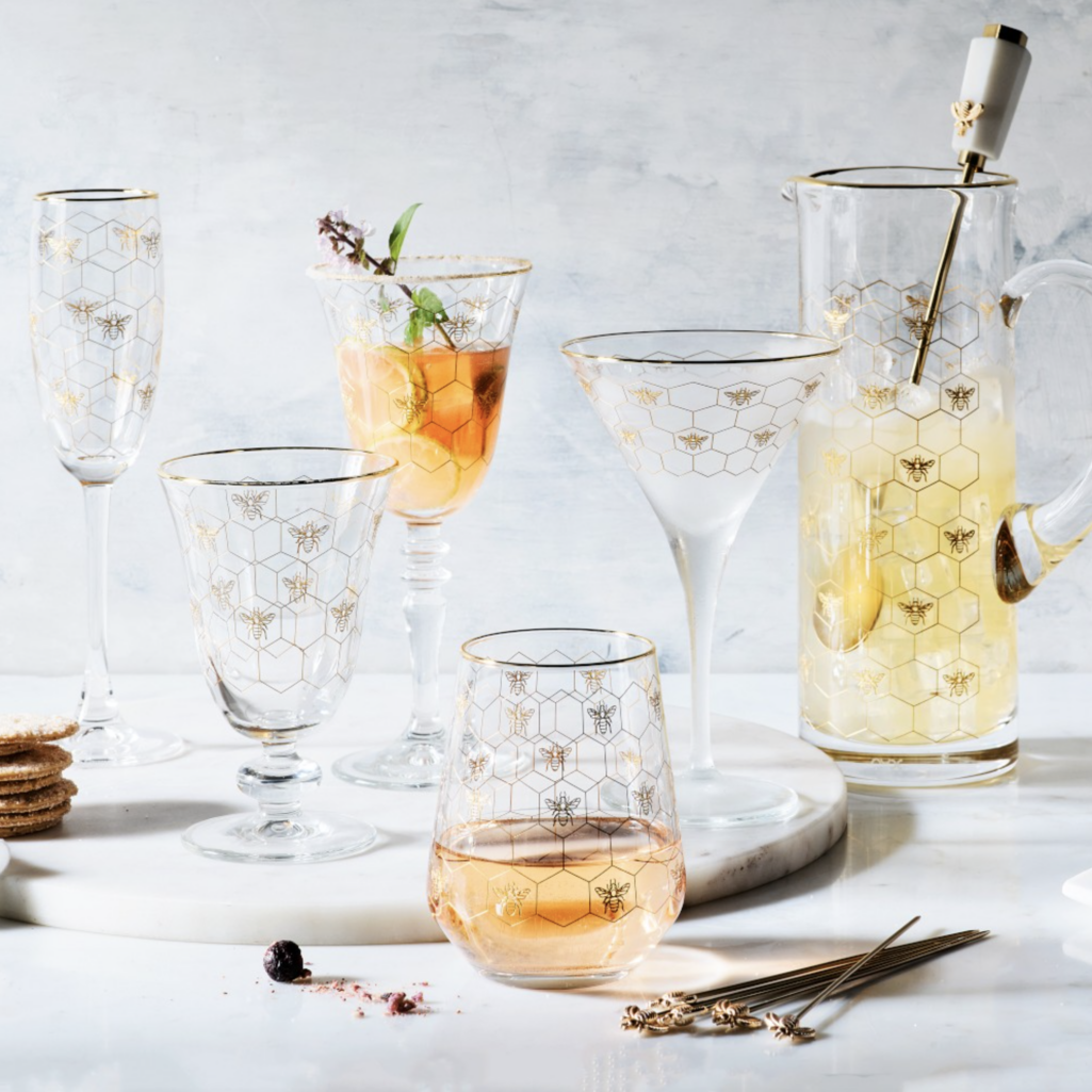 Assorted clear glassware with gold bee decals, honeycomb pattern, and gold rims, including wine glasses, stemless glasses, martini glasses, flutes, and a pitcher arranged on a tabletop.