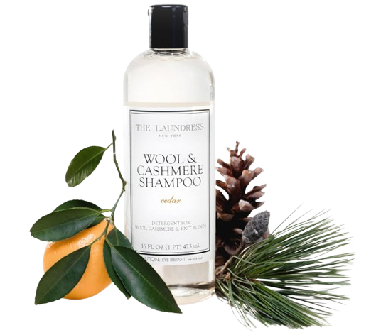 Clear bottle of The Laundress wool and cashmere shampoo with a black cap, styled with pinecones, greenery, and citrus to highlight its cedar scent.