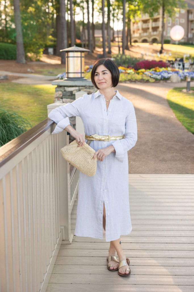 Kelly in pale blue linen shirtdress with gold braided belt, gold slide sandals and straw clutch on outdoor walkway — petite shirtdress evening styling