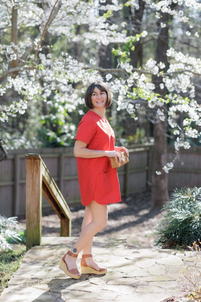 Side profile of Kelly in red shift dress with raffia wedge sandals and straw bee clutch, cherry blossom trees — petite spring dress silhouette