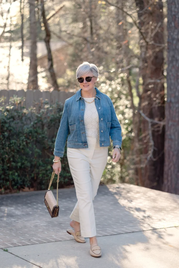 Woman over 50 with silver hair walking in an all-cream outfit — a lace tank layered under a cropped denim jacket with kick flare ivory pants and nude loafers, accessorized with a tortoise chain clutch, tortoise sunglasses, and gold hoop earrings — demonstrating how to wear soft white as a tonal spring look over 50.