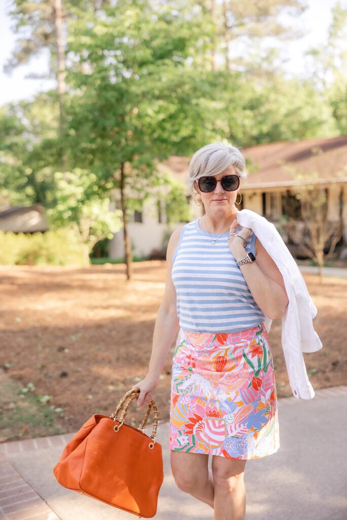 Woman over 50 with silver hair walking outdoors wearing a blue and white horizontal striped sleeveless tank top paired with a vibrant multicolor tropical floral mini skirt in pink, orange, lavender, and green, carrying a white denim jacket casually over one shoulder and an cognac leather tote with bamboo handles, styled with gold hoop earrings and dark sunglasses — an additional example of how to mix a classic stripe with a bold floral print over 50.