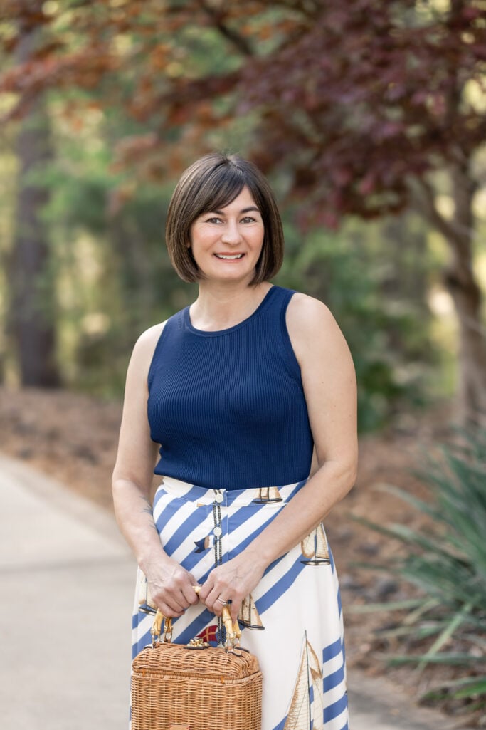 Three-quarter shot of Kelly in navy ribbed knit top tucked into Ralph Lauren sailboat stripe midi skirt with wicker bamboo bag — nautical petite outfit spring