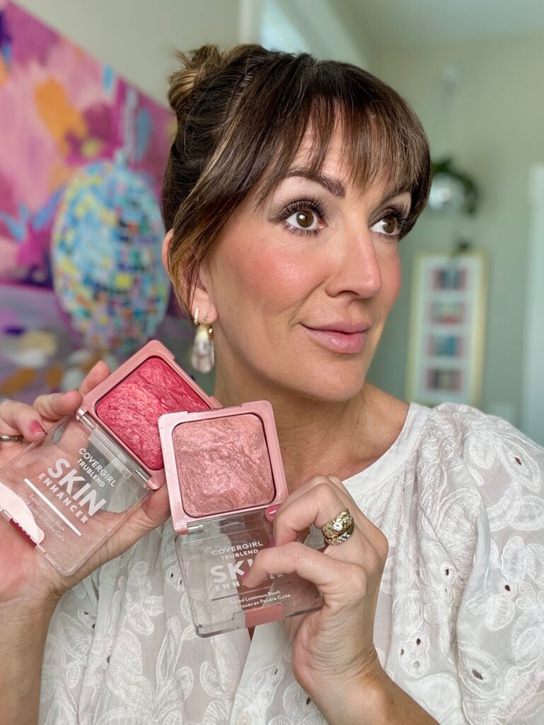 Close-up of woman making kissing lips expression holding Violette_Fr Bisou Balm white and gold tube in one hand and gold cap in other, showing blurred rosy pink lip result, wearing white eyelet top, clear lucite drop earrings with gold accent, pink manicure, completed blurred soft focus makeup look with rosy cheeks and taupe smudged eye, colorful disco ball painting in background