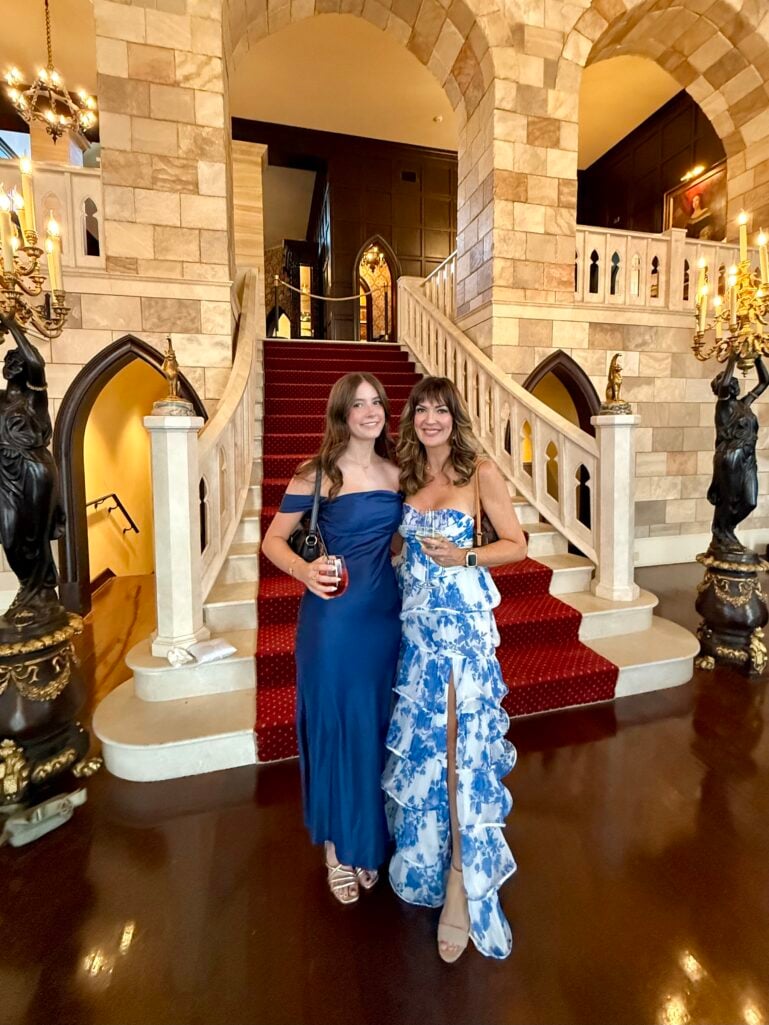 Woman holding flat-top kabuki facial brush with bamboo handle alongside Bondi Sands Self Tanning Foam bottle in Light/Medium shade, wearing white eyelet blouse, gold coil bangles and silver watch bracelet, clear drop earrings, no makeup fresh face with naturally bronzed skin, disco ball and hanging plant visible in background