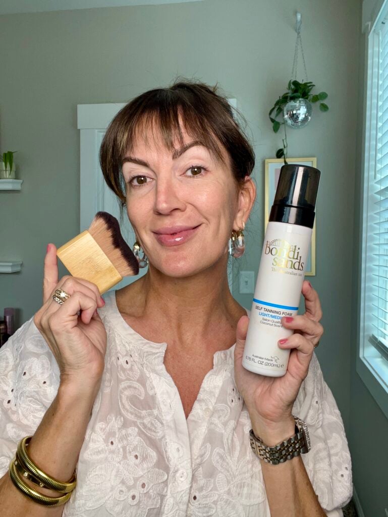 Woman holding open IT Cosmetics Do It All Sheer Tint Face Balm pot showing golden-toned balm formula alongside silver foundation buffing brush near cheek, wearing white eyelet blouse, clear lucite drop earrings, stacking gold rings, pink manicure, colorful abstract painting and disco ball in background