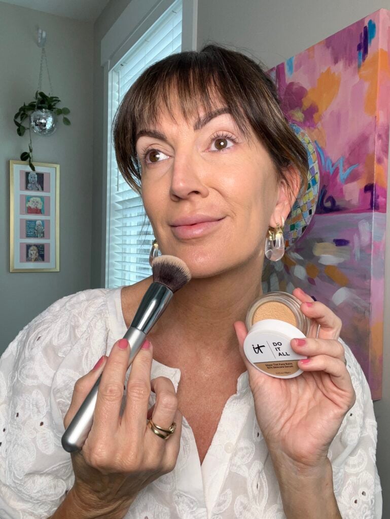 Woman holding NYX Professional Makeup Bare With Me Concealer Serum pink pump bottle while pointing to concealer dot applied under eye, wearing white eyelet blouse, clear lucite drop earrings, stacking rings, pink manicure, colorful abstract painting in background