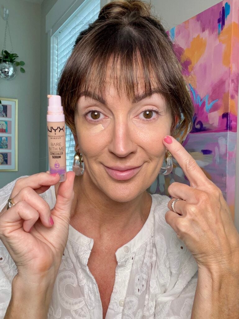 Close-up of hand holding two L'Oreal Paris Le Shadow Stick eyeshadow crayons — one in champagne shimmer and one in warm taupe brown shade labeled Celestial Taupe — pink manicure visible, mirror reflection and makeup products softly blurred in background