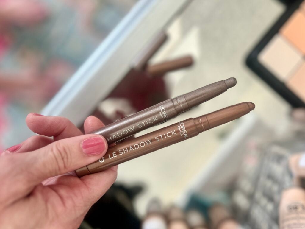 Close-up of hand holding two L'Oreal Paris Le Shadow Stick eyeshadow crayons — one in champagne shimmer and one in warm taupe brown shade labeled Celestial Taupe — pink manicure visible, mirror reflection and makeup products softly blurred in background