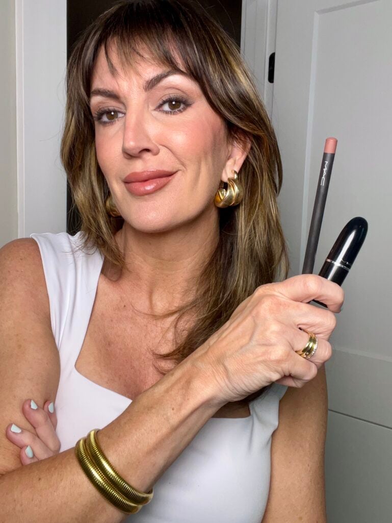 Woman holding MAC Cosmetics Spice lip liner pencil and closed MAC Lustreglass lipstick bullet together while wearing completed 90s-inspired makeup look with matte bronzed skin, brown smoky eye and warm nude-pink lips, white sleeveless top, gold chunky hoop earrings and gold coil bangles