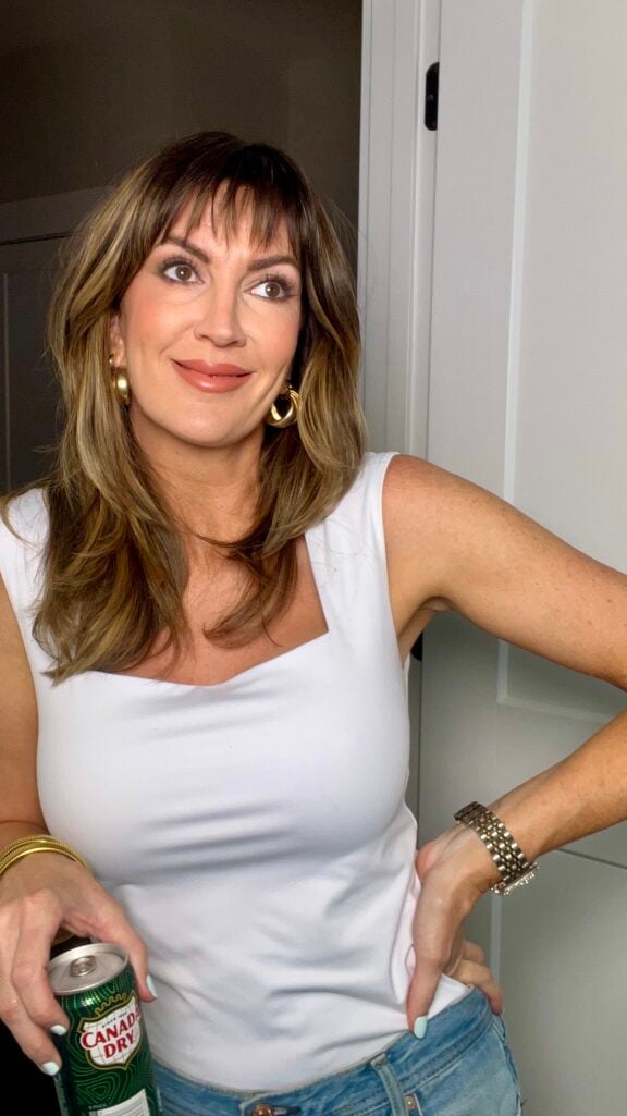 Woman in white square neck sleeveless top and light wash jeans posing with hand on hip holding Canada Dry ginger ale can at side in Cindy Crawford Pepsi ad-inspired pose, wearing gold chunky hoop earrings and gold coil bangles, completed 90s makeup look with matte skin and nude-pink lips, silver watch bracelet on opposite wrist