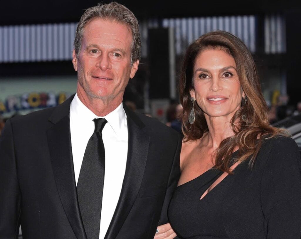 Man in black suit with black tie and woman in black asymmetric one-shoulder gown with silver chandelier earrings posing together at formal event, woman with long highlighted brunette waves showing naturally radiant and youthful aging skin