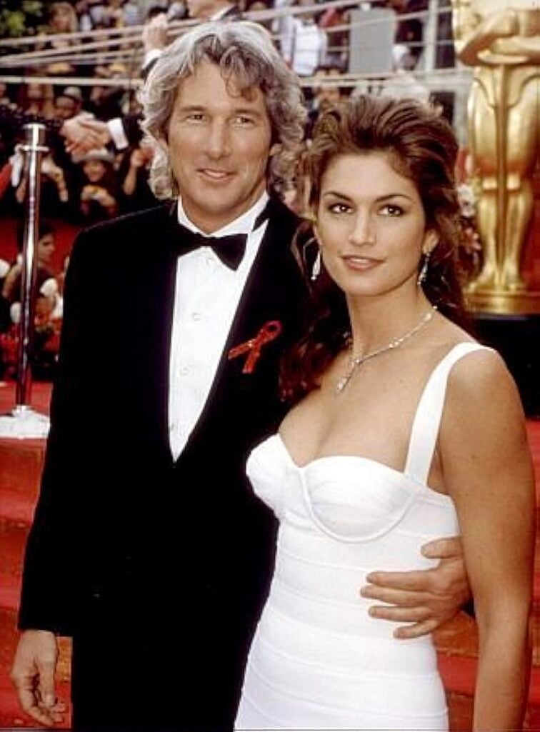 Vintage 1990s red carpet photo of man in black tuxedo with red ribbon pin and woman in white fitted bustier gown with upswept voluminous dark hair, pearl drop earrings and diamond necklace, gold Oscar statuettes visible in background
