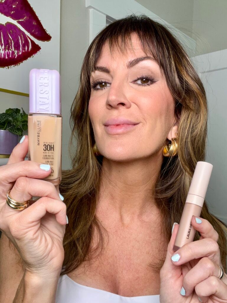 Woman holding Maybelline SuperStay 30H Lumi Matte Foundation bottle in shade 125 in left hand and Laura Mercier Real Flawless Weightless Perfecting Concealer tube in right hand, wearing white square neck top, gold bubble hoop earrings, stacking rings, mint blue manicure, matte base makeup look