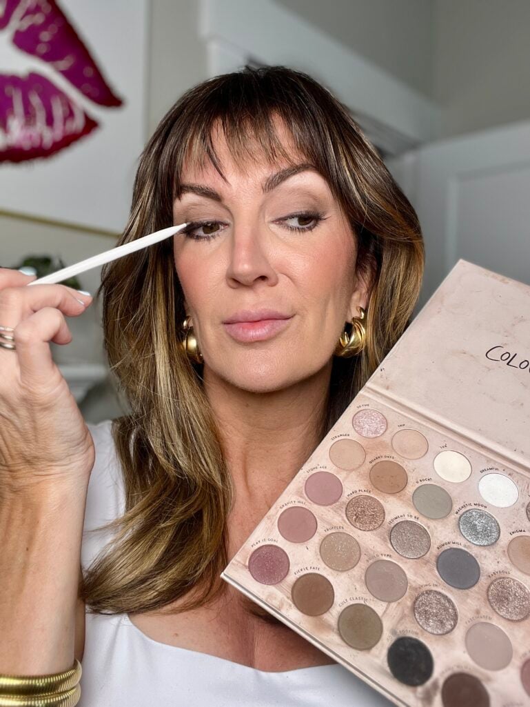 Woman applying eyeshadow with small white liner brush to upper lash line while holding open Colourpop Stone Cold Fox neutral eyeshadow palette showing shades including Stranger, Quarry Days, Hard Place, Truffles and more, wearing white square neck top and gold chunky hoop earrings and gold coil bangles, pink lip wall art in background