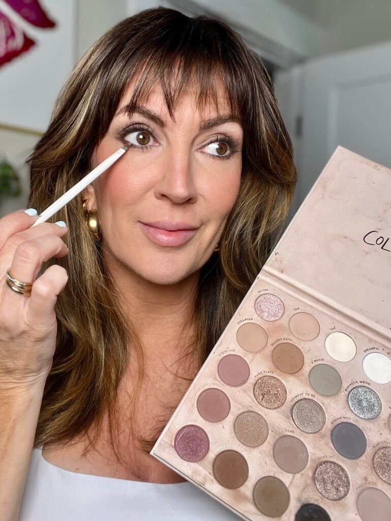 Woman smiling and looking up while holding open Colourpop Stone Cold Fox palette showing full range of warm neutral and taupe matte and shimmer eyeshadows, applying shadow with white liner brush to lower lash line, wearing white top, gold bubble hoops, stacking rings and gold coil bangles, mint blue nails