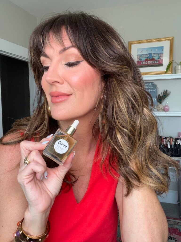 Woman in red V-neck top with eyes closed smiling contentedly while holding Gisou Honey Infused Hair Oil dropper bottle with gold cap, showing glossy highlighted brunette waves, gold hoop earrings, jeweled gold cuff bracelet, stacking rings with red cherry nail art, Cotton Candy Sno-Kones framed art and makeup brush collection visible on shelf in background