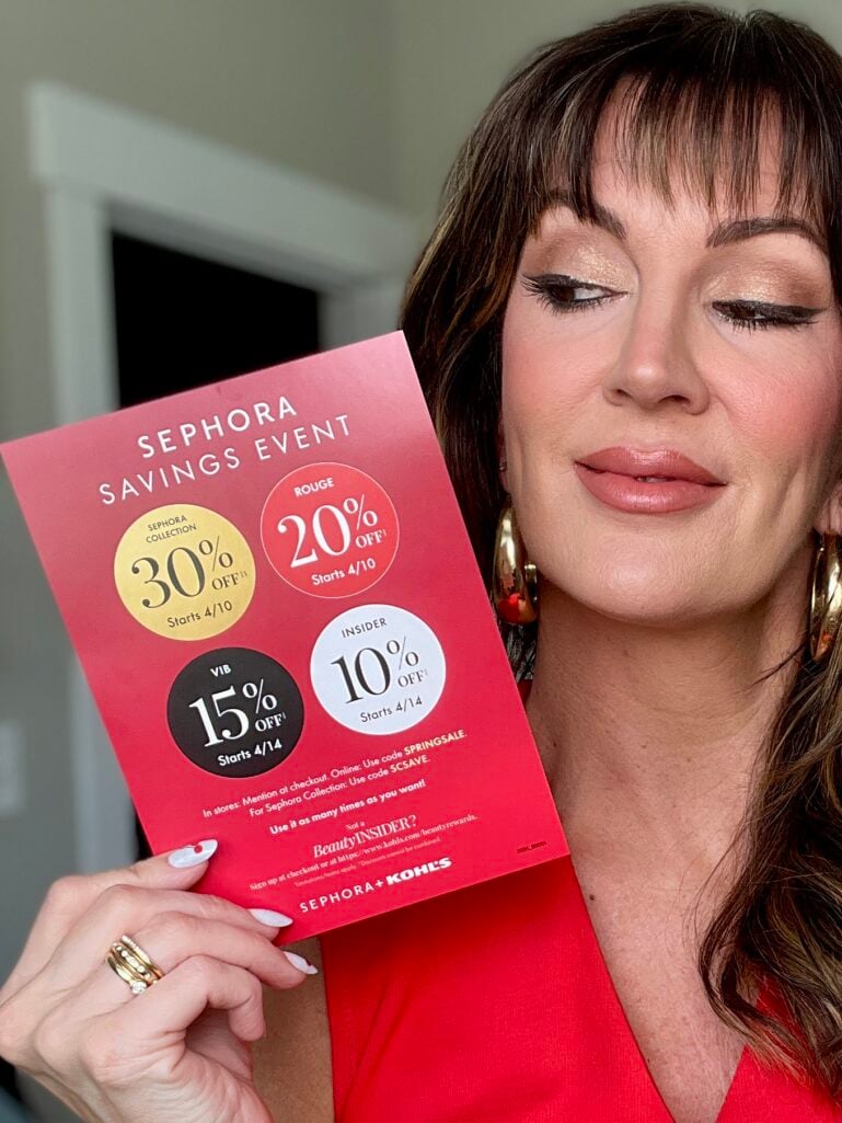 Close-up of woman in red V-neck top holding red Sephora Savings Event card showing Rouge 20% off starting 4/10, VIB 15% off and Insider 10% off starting 4/14, gold chunky hoop earrings visible, warm bronzed eye makeup with dramatic liner, stacking gold rings, white and red cherry nail art