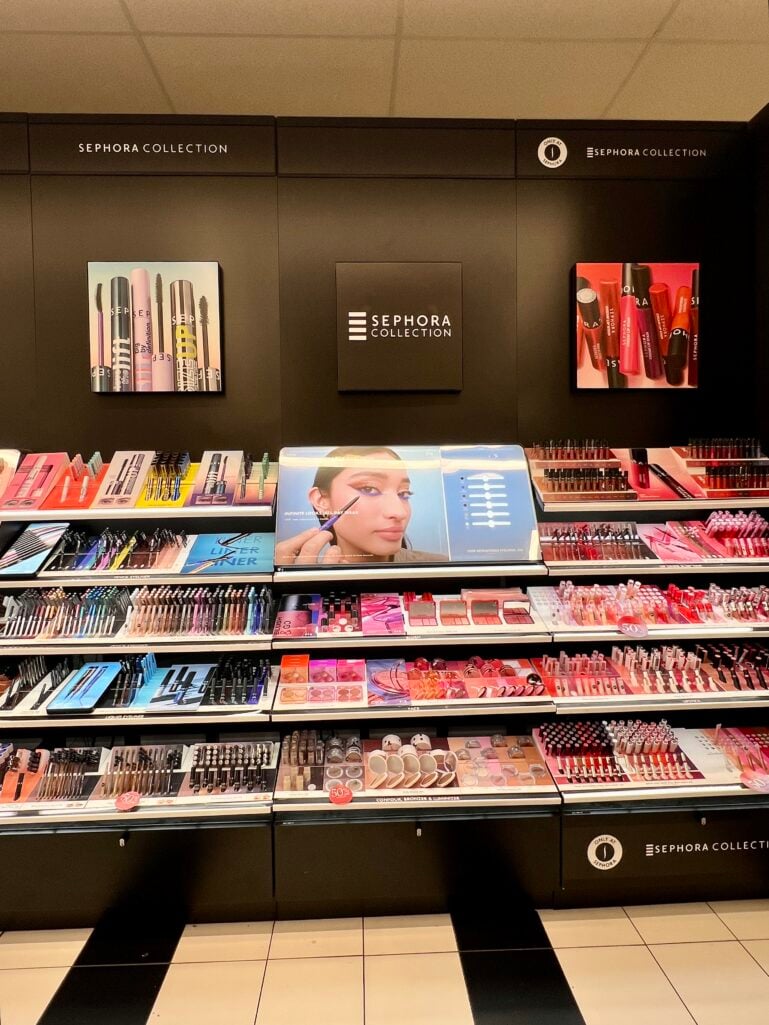 Inside Sephora Collection display wall at Kohl's showing black shelving units filled with colorful makeup products including mascaras, eyeliners, blushes, lip colors and contour products, illuminated signage reading Sephora Collection, promotional imagery featuring model with graphic blue liner