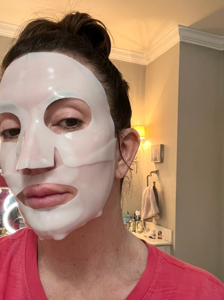 Woman wearing white Biodance Collagen Real Deep Sheet Mask covering entire face with cutouts for eyes, nose and mouth, hair pulled back loosely, pink top, bathroom visible in warm lamplight background