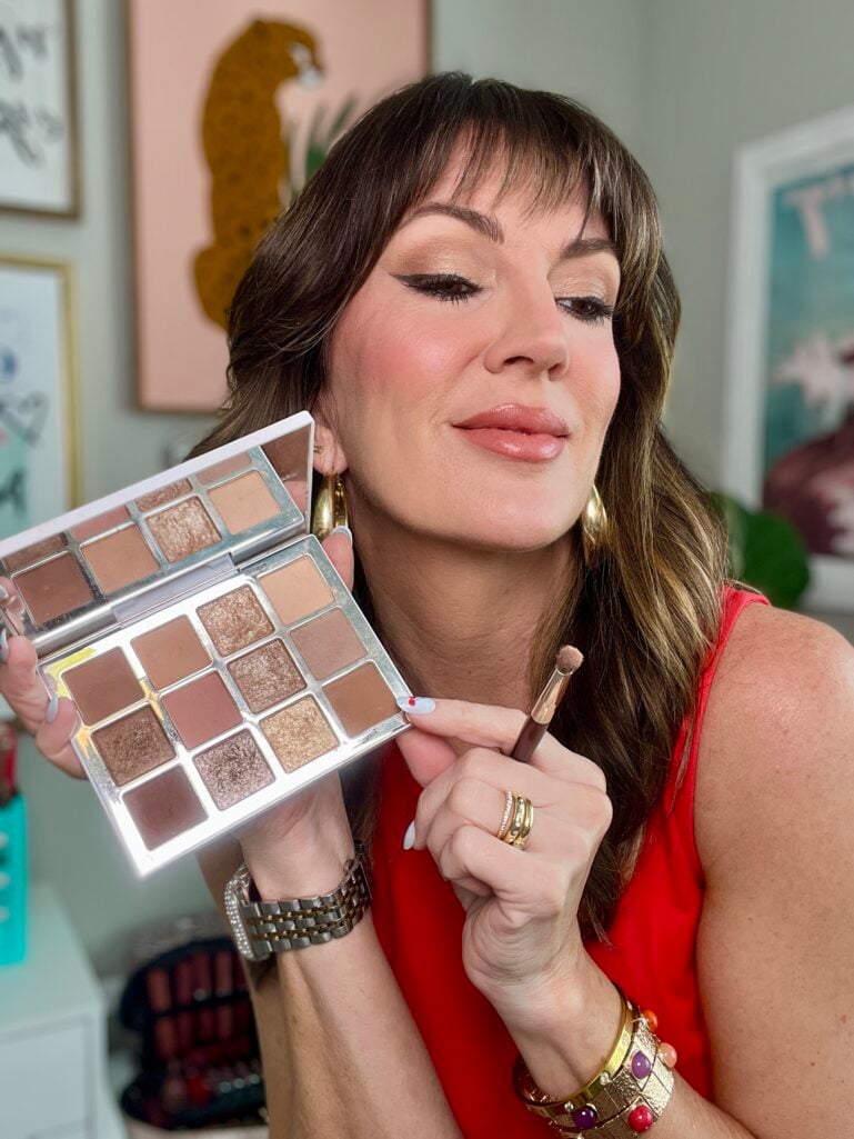 Woman in red top looking forward smiling while holding open Makeup by Mario Ethereal Eyes eyeshadow palette displaying warm brown and champagne neutral shades, holding small eyeshadow blending brush, gold chunky hoop earrings, stacking gold rings, silver watch bracelet, white and red nail art, botanical print artwork visible in background