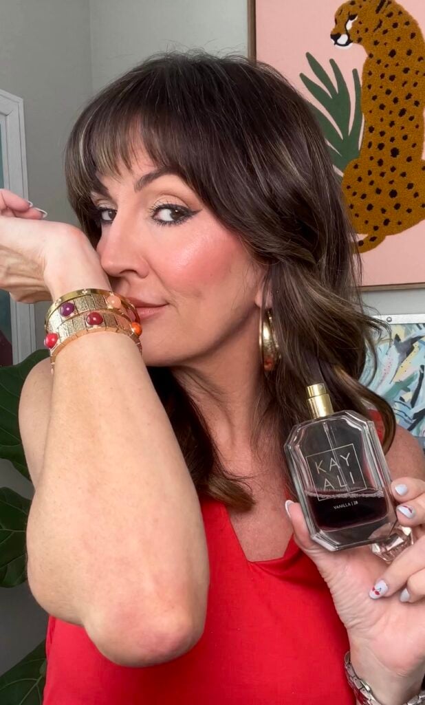 Woman in red V-neck top posing with head resting on jeweled gold cuff bracelet wrist while holding Kayali Vanilla 28 dark amber glass perfume bottle with gold spray cap, wearing gold chunky hoop earrings, looking sideways with soft smile, cheetah print art and tropical leaf botanical print visible in background