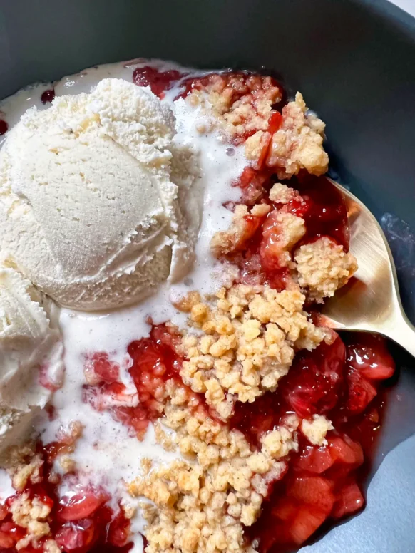 Close-up of a serving of skillet strawberry-rhubarb crisp in a slate blue bowl with a scoop of melting vanilla ice cream and a gold spoon, showing the jammy red fruit filling and golden buttery panko topping