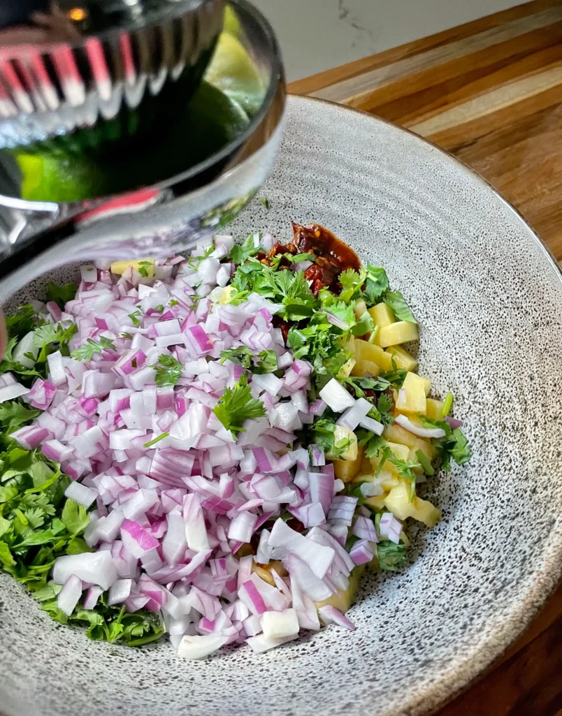 Chipotle pineapple salsa ingredients being combined in a speckled ceramic bowl: diced red onion, fresh cilantro, diced pineapple, and chipotle peppers in adobo sauce, with a lime juicer in the background