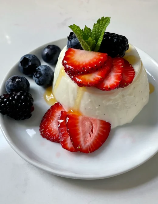 Close-up of an unmolded buttermilk vanilla panna cotta on a white plate, topped with sliced strawberries, blueberries, blackberries, and fresh mint with a drizzle of honey