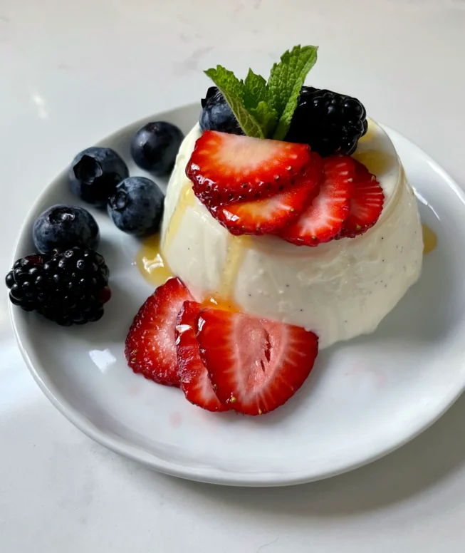 Close-up of an unmolded buttermilk vanilla panna cotta on a white plate, topped with sliced strawberries, blueberries, blackberries, and fresh mint with a drizzle of honey