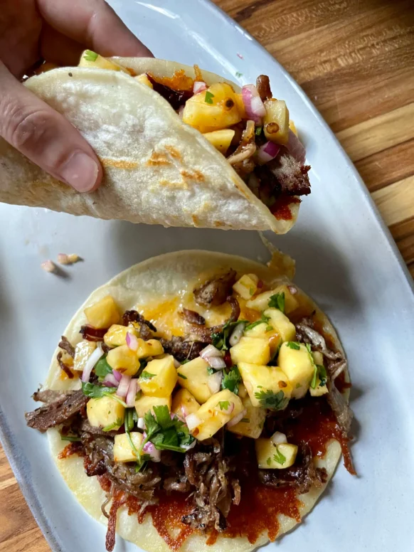 Hand holding a folded pork taco with chipotle pineapple salsa over a white platter with a second assembled taco, showing the crispy cheese underside of the tortilla