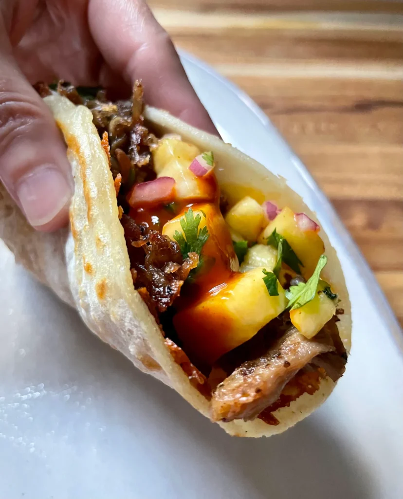 Close-up of a hand holding a folded pork taco showing the crispy cheese layer, shredded pork, chipotle pineapple salsa, and hot sauce inside a corn tortilla
