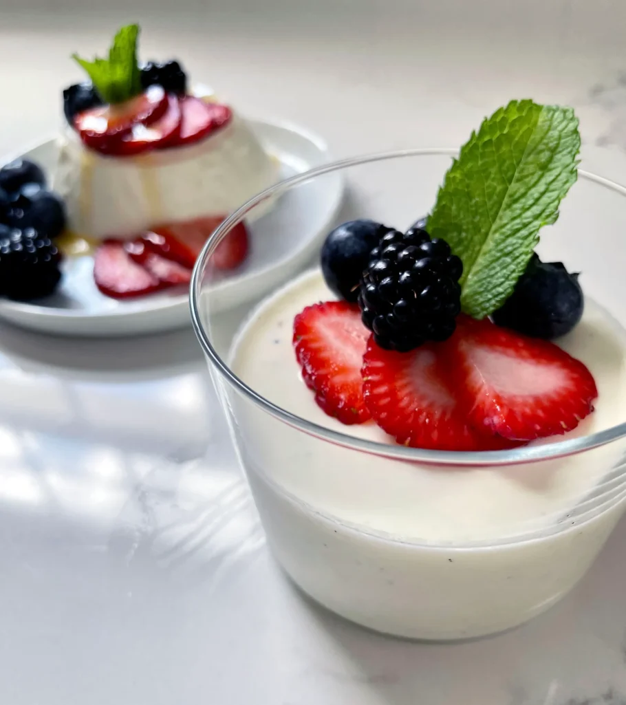 Angled shot of a buttermilk vanilla panna cotta served in a clear glass cup in the foreground topped with strawberries, a blackberry, blueberries, and a mint sprig, with an unmolded plated panna cotta visible in the background