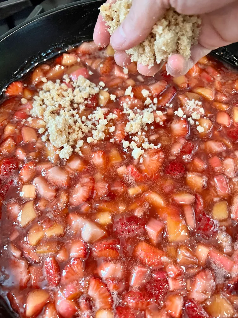 Hand crumbling and sprinkling buttery panko crisp topping over the hot strawberry-rhubarb filling in a cast iron skillet