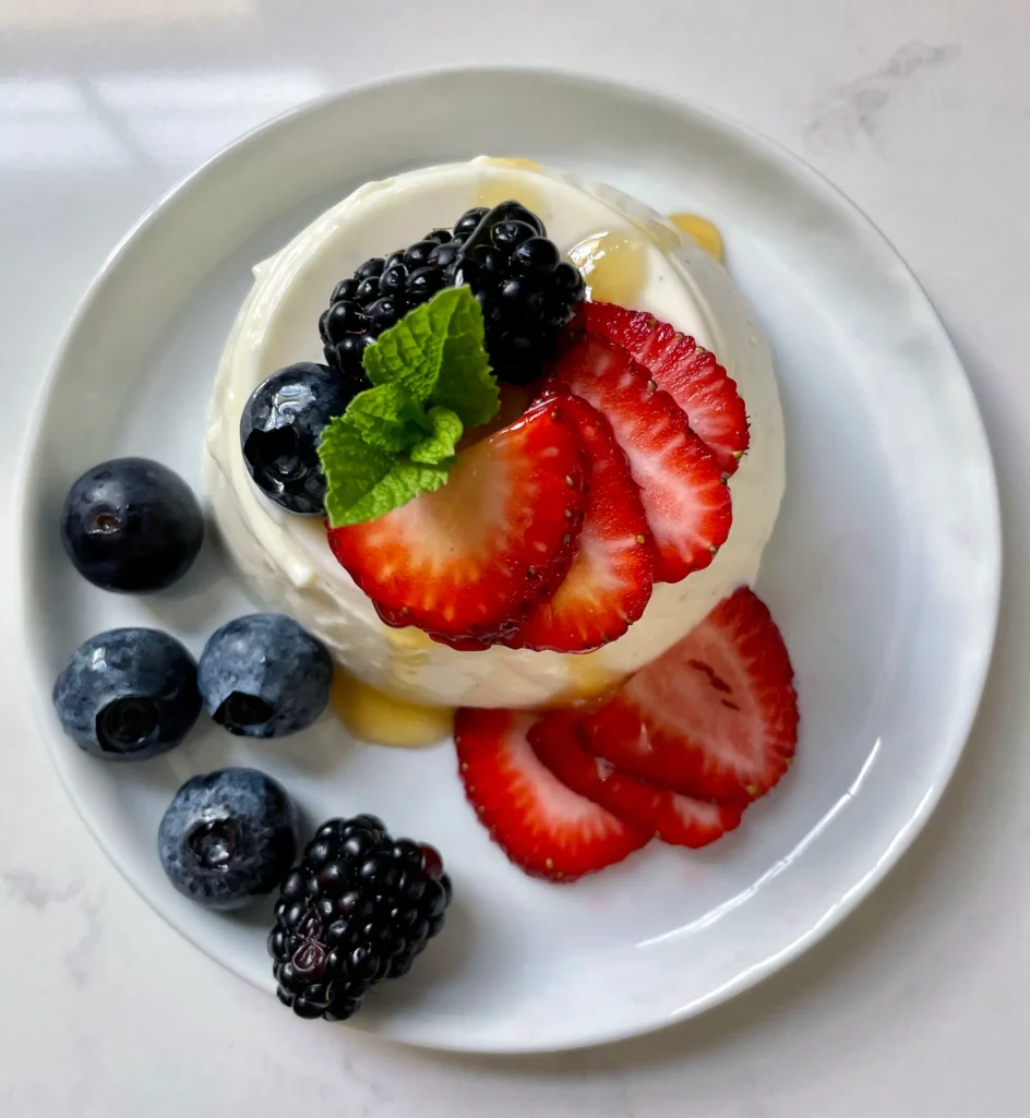 Overhead view of an unmolded buttermilk vanilla panna cotta on a white plate topped with sliced strawberries, blackberries, blueberries, and fresh mint with a honey drizzle, surrounded by additional fresh berries on the plate