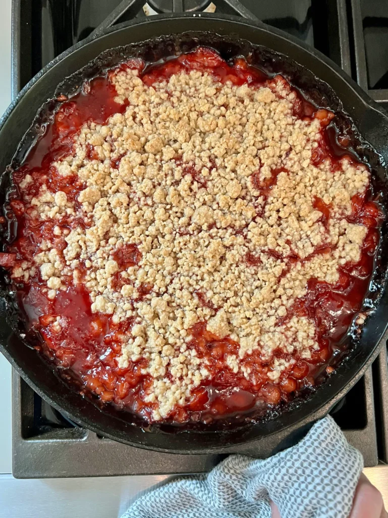Freshly baked skillet strawberry-rhubarb crisp in a cast iron skillet, with golden panko topping and deep red fruit filling bubbling up around the edges, held with a kitchen towel