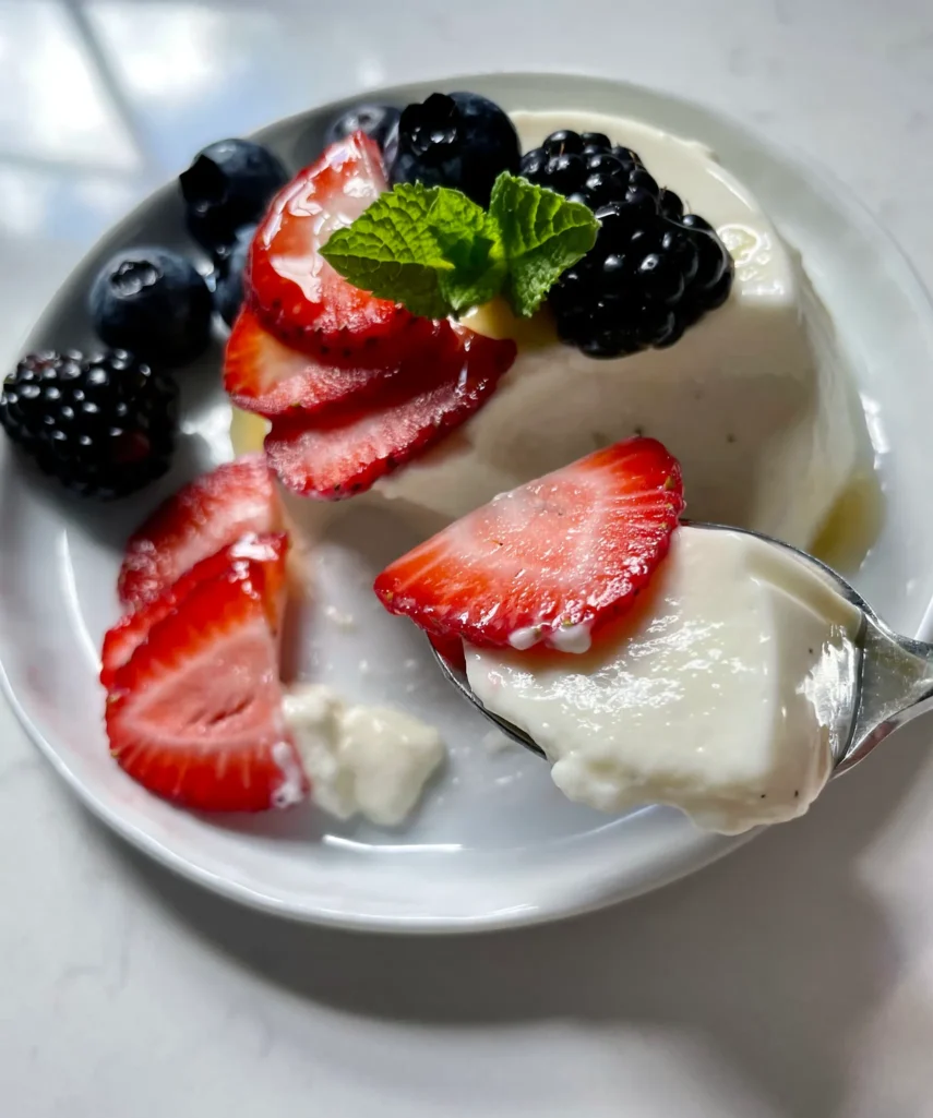 Close-up of a spoonful of silky buttermilk vanilla panna cotta being lifted from a plated serving alongside sliced strawberries, blackberries, blueberries, and fresh mint