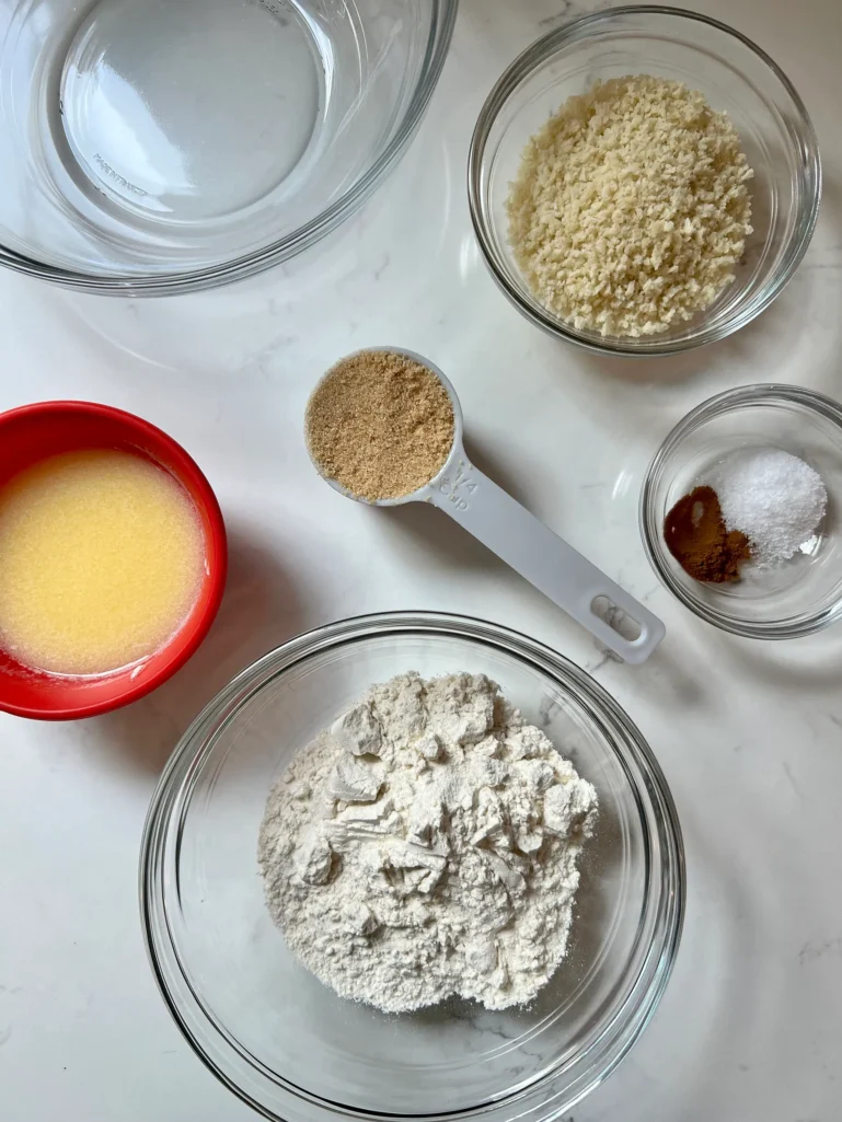 Overhead flat lay of crisp topping ingredients on a marble surface: a bowl of panko breadcrumbs, all-purpose flour, a measuring cup of brown sugar, a small bowl with salt and cinnamon, and a red bowl of melted butter