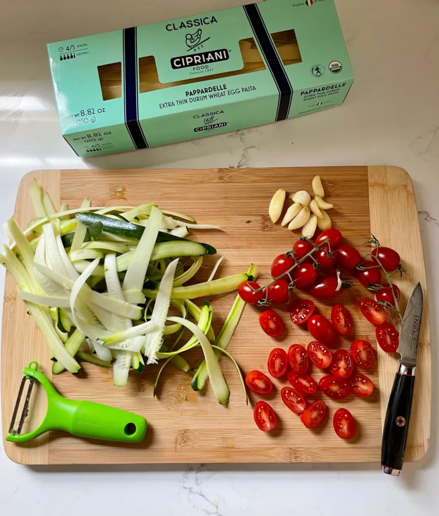 Flat lay of summer pasta ingredients on a wood cutting board: a box of Cipriani pappardelle, peeled zucchini ribbons, halved grape tomatoes on the vine, garlic cloves, a green vegetable peeler, and a paring knife
