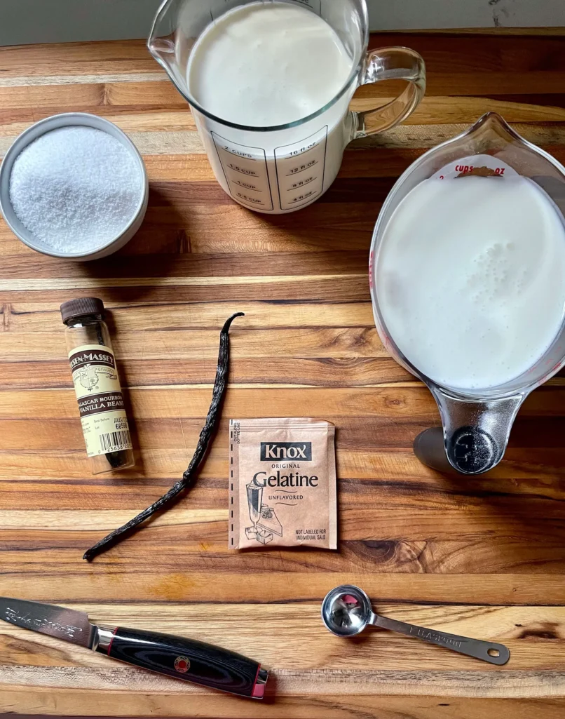 Flat lay of panna cotta ingredients on a wood cutting board: sugar in a tin, heavy cream in a large measuring cup, buttermilk in a batter bowl, a Nielsen-Massey vanilla bean, a Knox unflavored gelatin packet, a paring knife, and a teaspoon measure