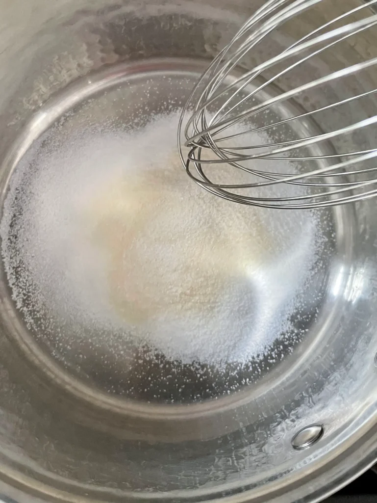 Overhead view of sugar, unflavored gelatin, and a pinch of salt being whisked together in a stainless saucepan before the cream is added