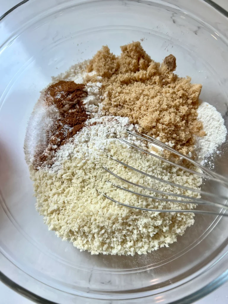 Overhead view of a glass mixing bowl with panko, all-purpose flour, brown sugar, cinnamon, and salt being whisked together to make the crisp topping