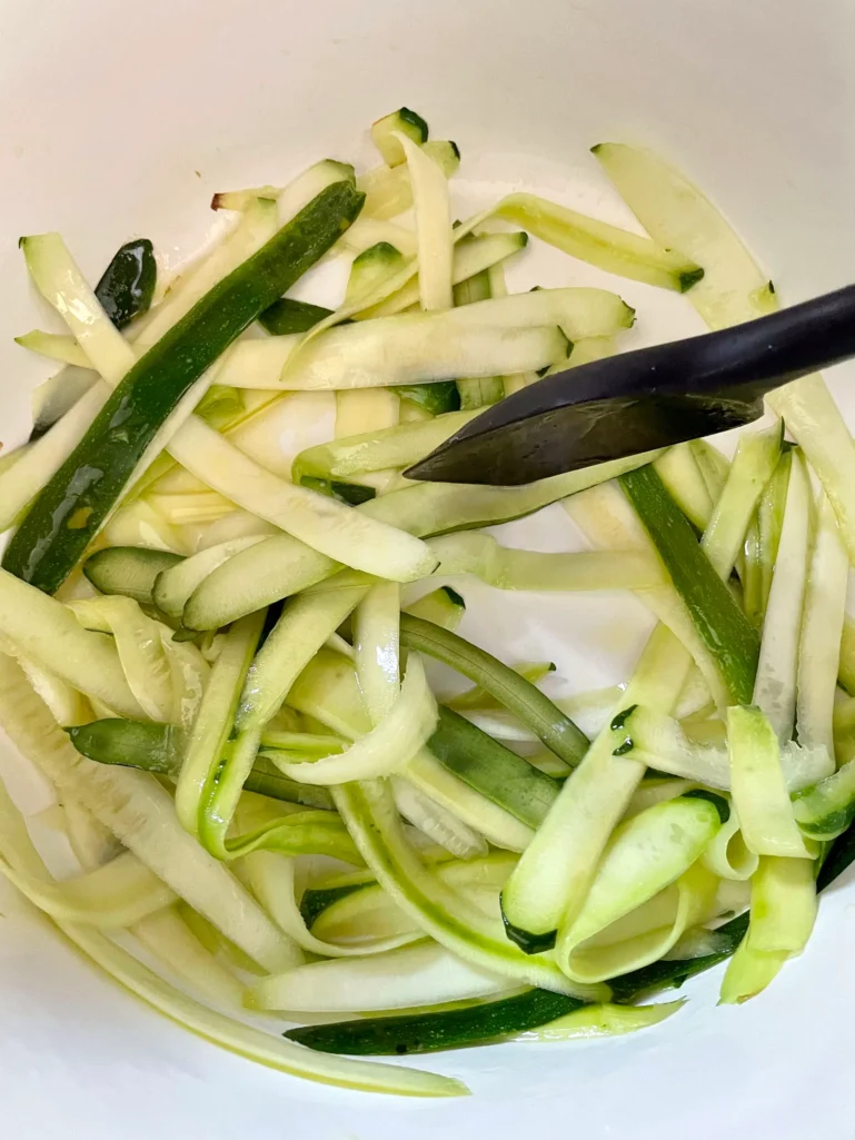Sautéed zucchini ribbons being tossed with black tongs in a large white Dutch oven, showing the thin peeler-cut strips turning slightly translucent