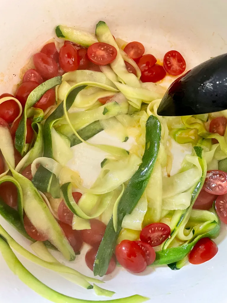 Halved grape tomatoes added to the softened zucchini ribbons in a white Dutch oven, beginning to release their juices with black tongs stirring the mixture