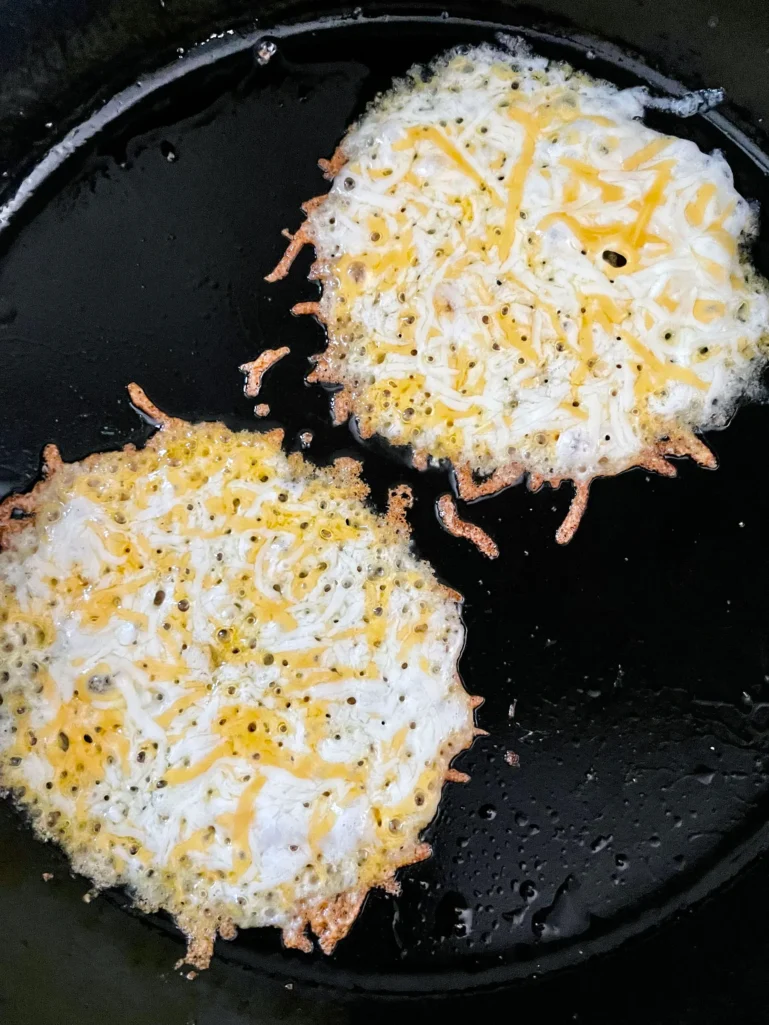 Two portions of shredded Mexican cheese blend bubbling and browning in a cast iron skillet, ready for the tortillas to be pressed on top