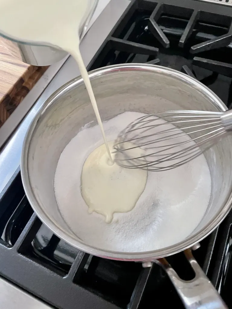 Heavy cream being poured from a glass measuring cup into the sugar and gelatin mixture in a saucepan on the stovetop
