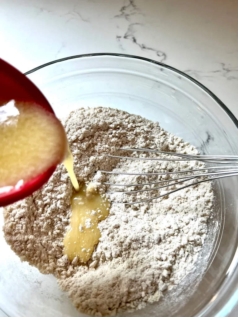 Melted butter being poured from a red bowl into the whisked dry crisp topping ingredients in a glass mixing bowl