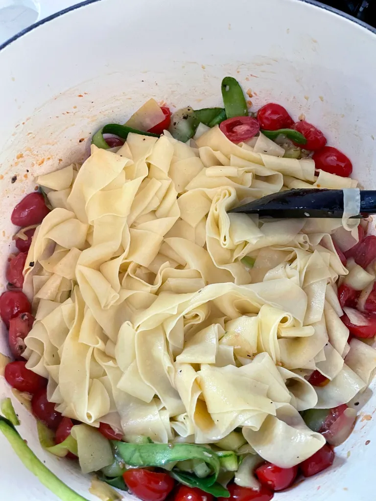 Hot cooked pappardelle being added directly on top of the zucchini and tomato mixture in a white Dutch oven with black tongs