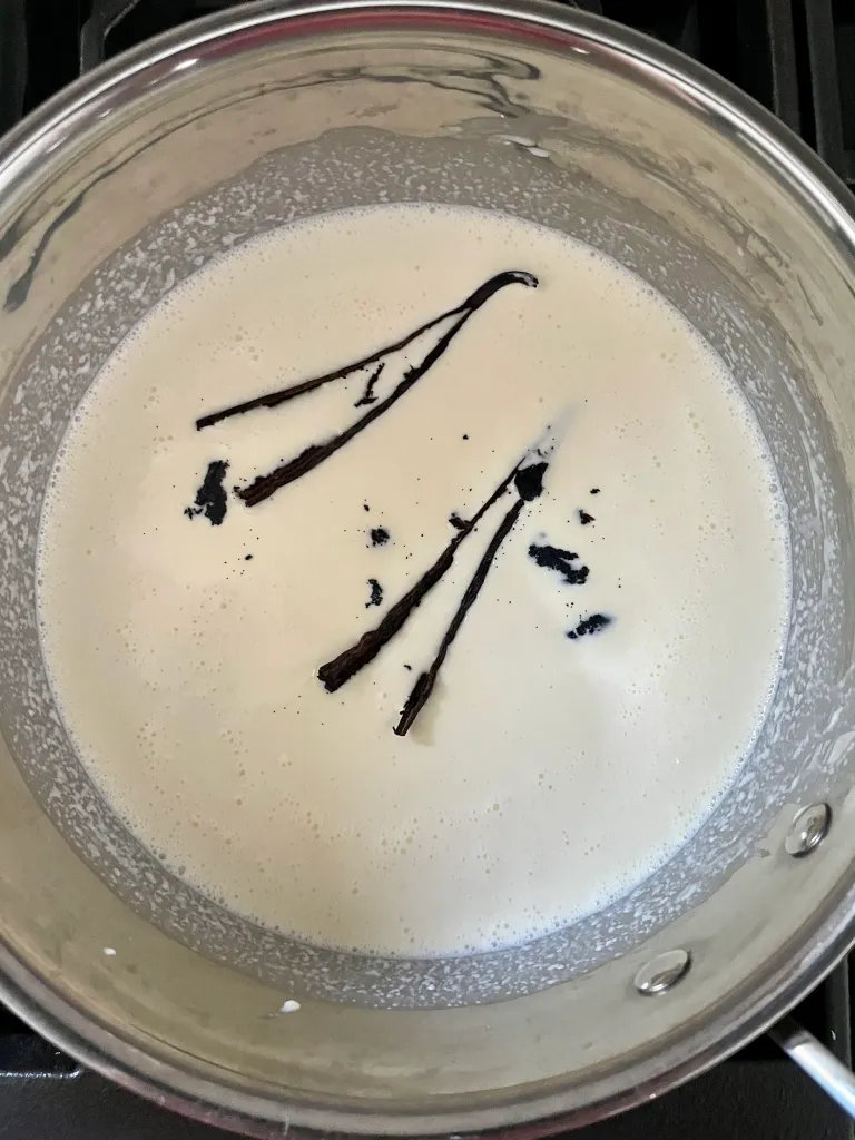 Overhead view of the cream mixture heating in a saucepan with split vanilla bean pods and scraped vanilla seeds steeping in the cream