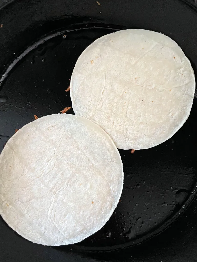 Two plain corn tortillas toasting in a cast iron skillet after being flipped onto the melted cheese